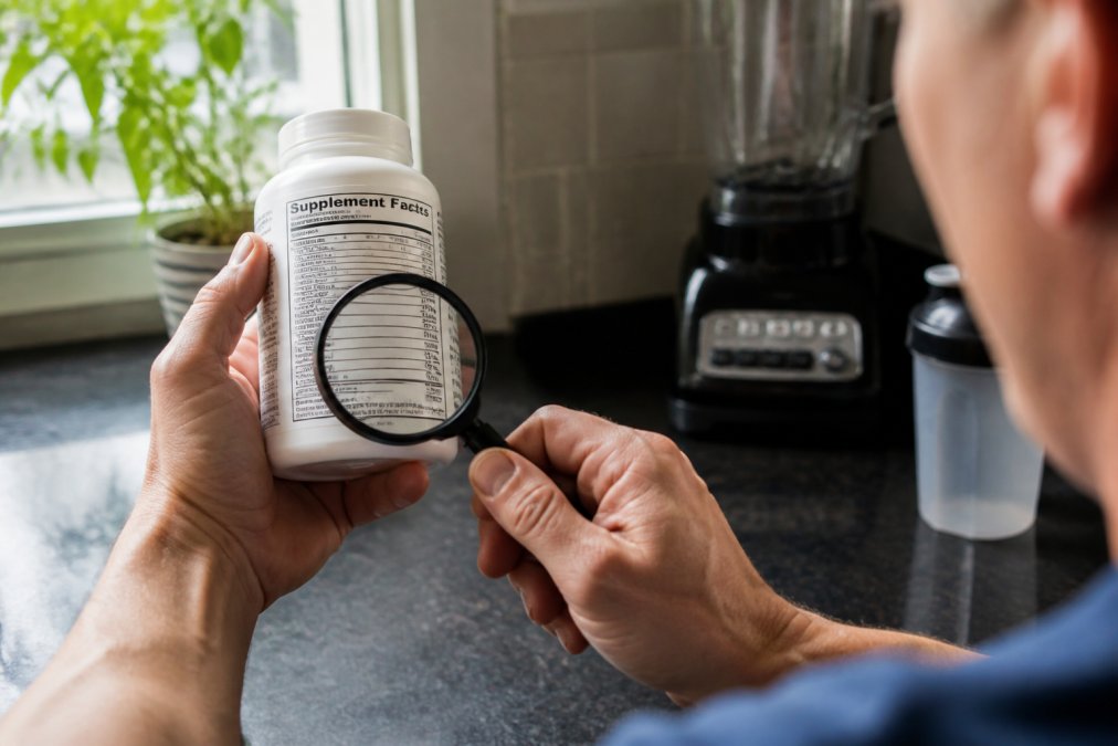 Close-up of a person's hands holding a supplement bottle, with the Supplement Facts panel clearly visible and in focus, a magnifying glass resting on the label suggesting close inspection, natural kitchen lighting