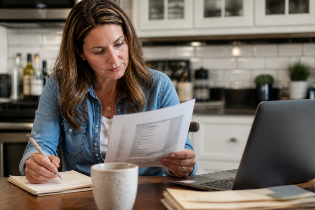 A person sitting at a kitchen table with a printed lab report in one hand and a pen in the other, reading the antibody panel section closely, with a laptop open nearby showing a patient portal