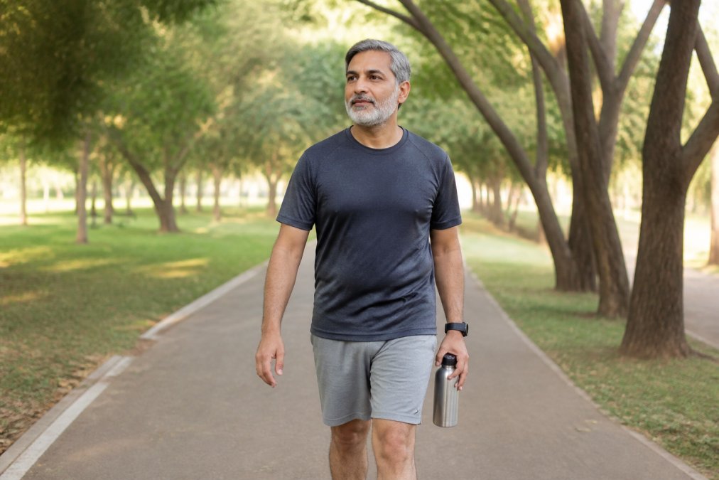 A middle-aged adult walking outdoors on a paved path in a quiet park, wearing comfortable athletic clothing and holding a stainless steel water bottle, with a calm and determined expression