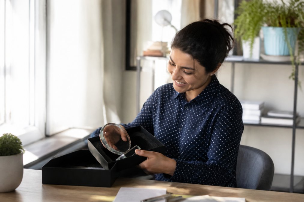 Remote employee smiling at their home desk while carefully opening a personalized service award package, with the award visible and natural window lighting illuminating their face