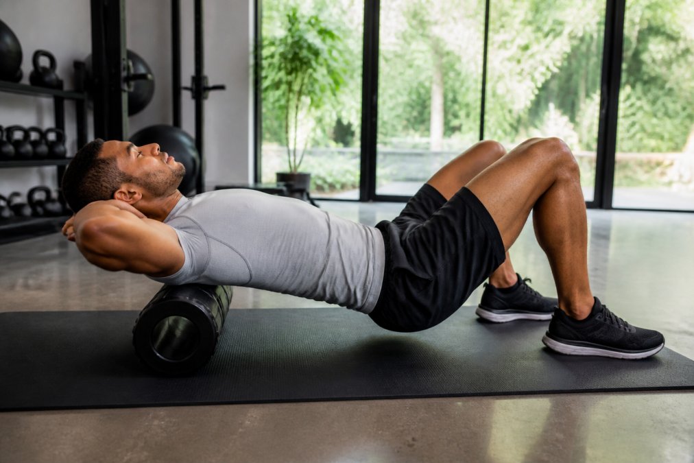 A person using a foam roller on their upper back on a yoga mat in a bright, modern home gym, wearing athletic clothing, natural light coming through large windows, expression relaxed and focused