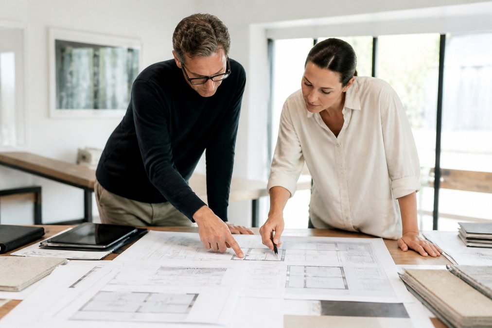 An architect and a building energy consultant reviewing residential floor plans and specification sheets spread across a desk in a bright design studio, pointing at details together