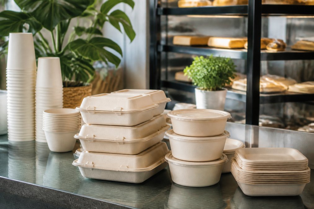 Stack of certified compostable takeaway containers and cups displayed on cafe counter with green sustainability signage and natural morning light