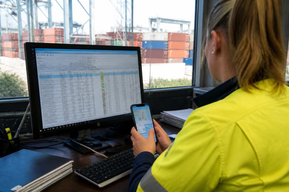 A logistics coordinator in a busy Australian port office checks container status on a large desktop monitor while holding a smartphone showing a tracking app, natural daylight through warehouse windows