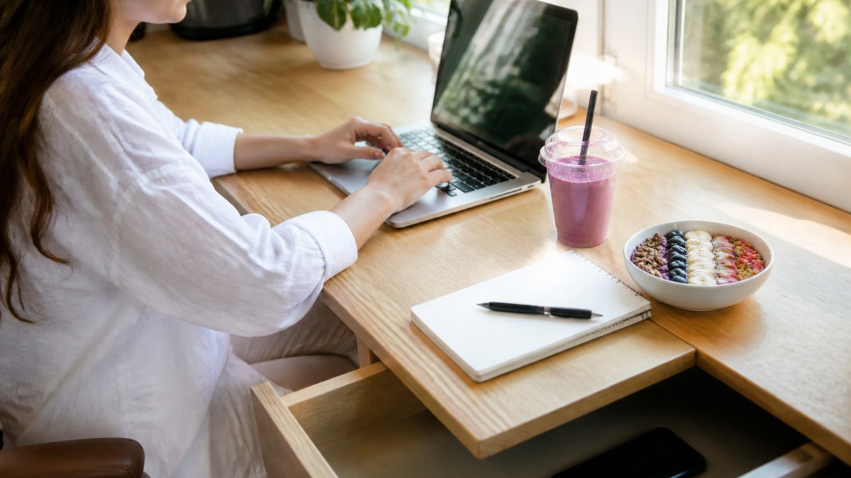 A woman at a tidy desk with her phone face-down in a drawer and a single laptop open, working in bright natural light with a smoothie and notebook beside her