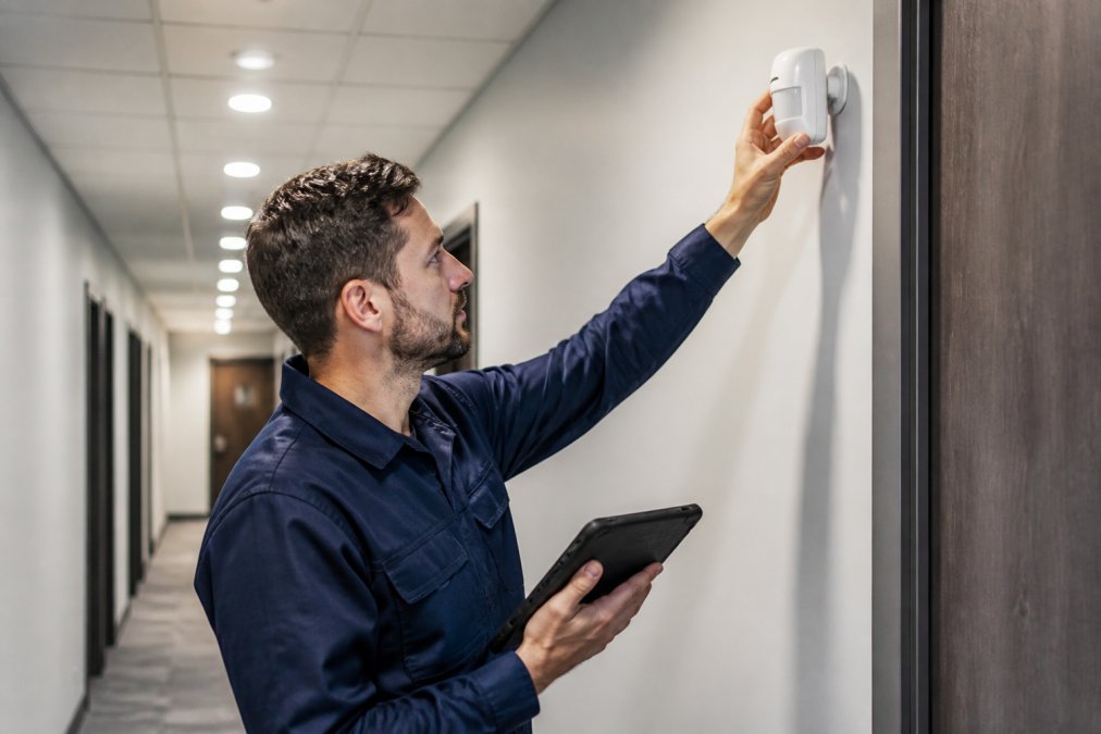 A security technician adjusting a wall-mounted motion detector in a modern commercial hallway, checking alignment with a tablet in hand under bright overhead lighting