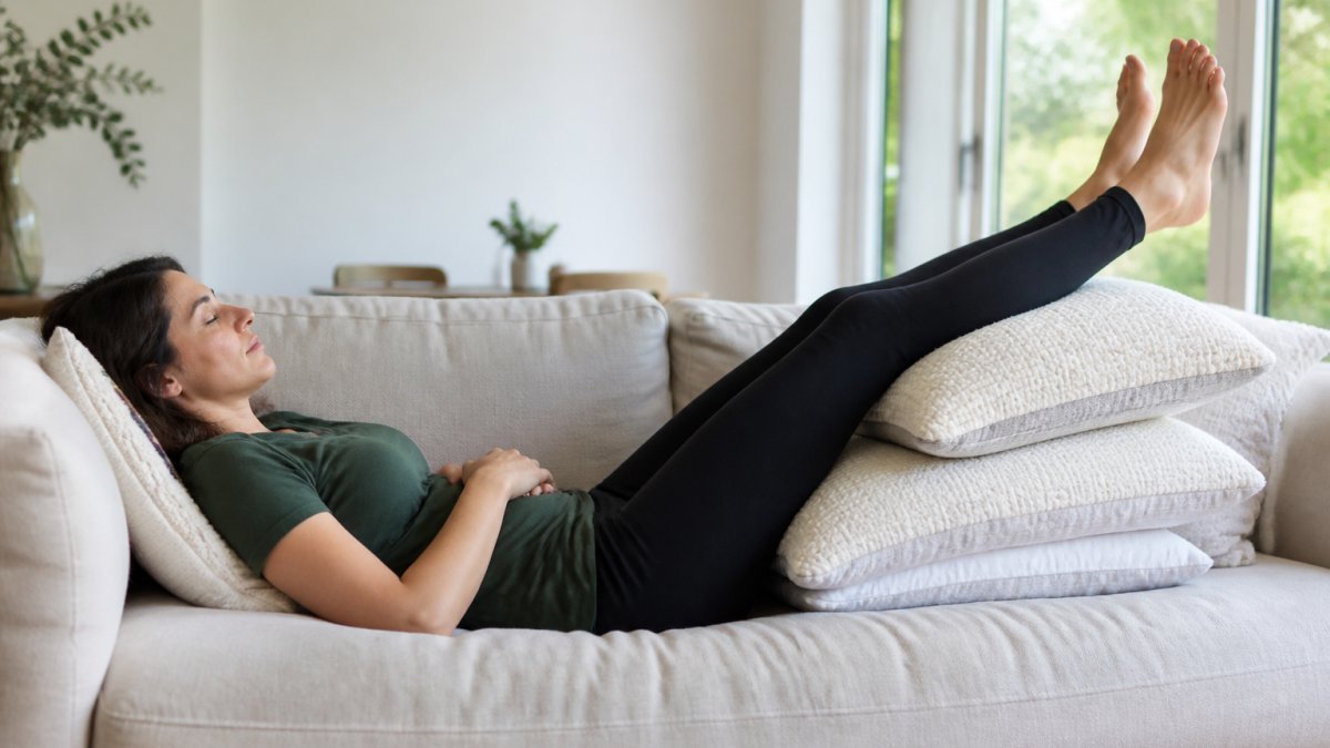 Person lying on a couch with legs elevated on pillows at approximately 45 degrees, demonstrating proper leg elevation for fluid retention relief in a bright, comfortable living room setting