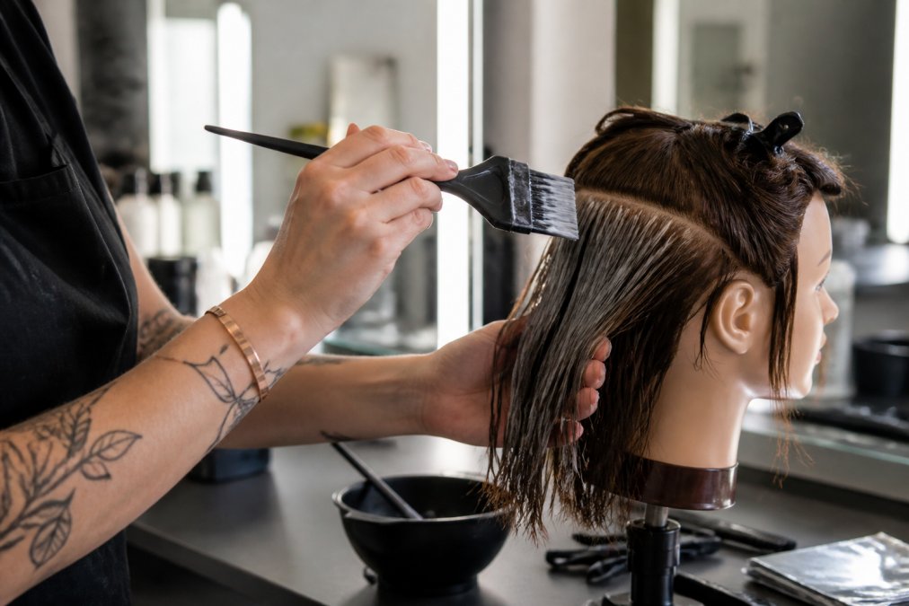 Close-up of a professional hair colorist's hand holding an ergonomic feathered tint brush with a relaxed, wide grip, applying color to a section of hair on a mannequin head in a bright salon