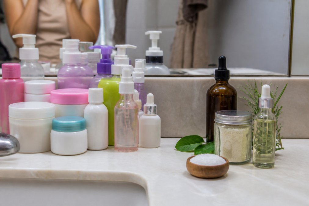 A woman's bathroom counter showing a mix of plastic containers, conventional beauty products, and a glass bottle with natural alternatives, illustrating the contrast between hormone-disrupting and safer product choices