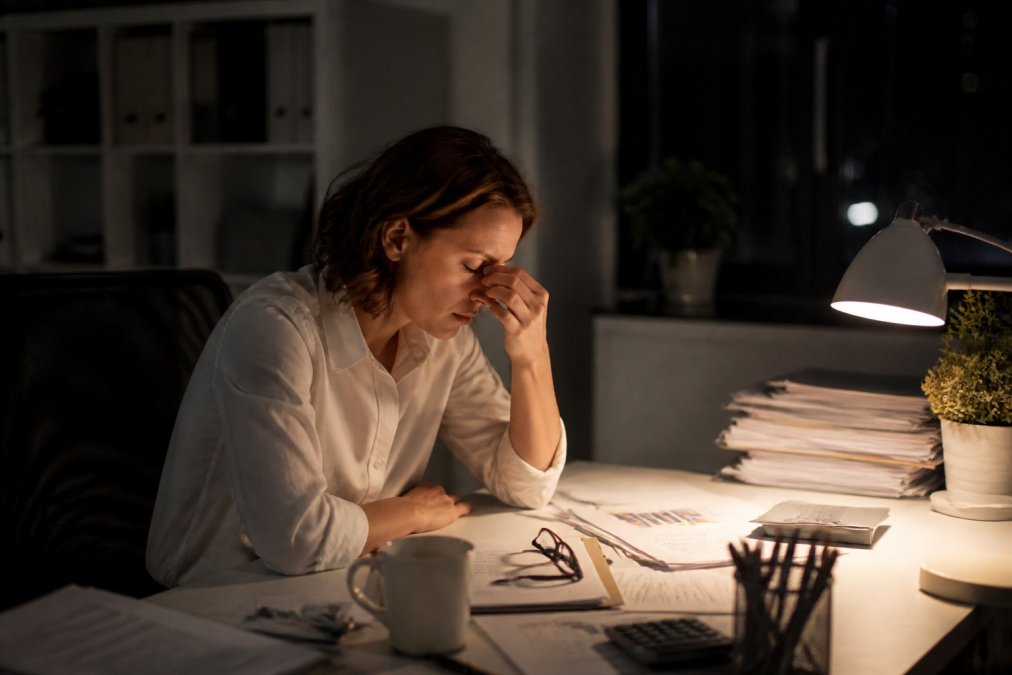 A manager sitting alone at a desk late in the evening, laptop open, papers spread out, looking tired and stressed in a dimly lit office