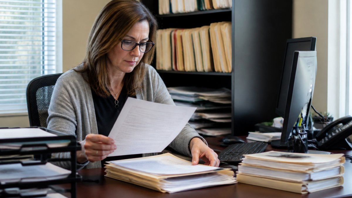 Practice manager reviewing provider enrollment documents at a desk with multiple files, computer screen showing credentialing dashboard, organized filing system in background