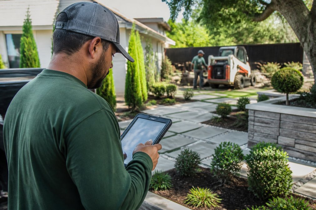 Field service technician on a job site using a mobile tablet to document completed work and send an invoice to the client, with finished landscaping and equipment visible in the background