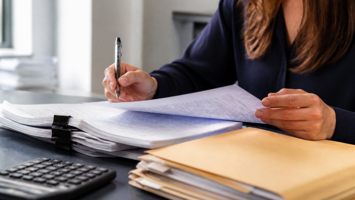 Practice manager at desk reviewing credentialing applications and insurance documentation with focused attention, natural office lighting