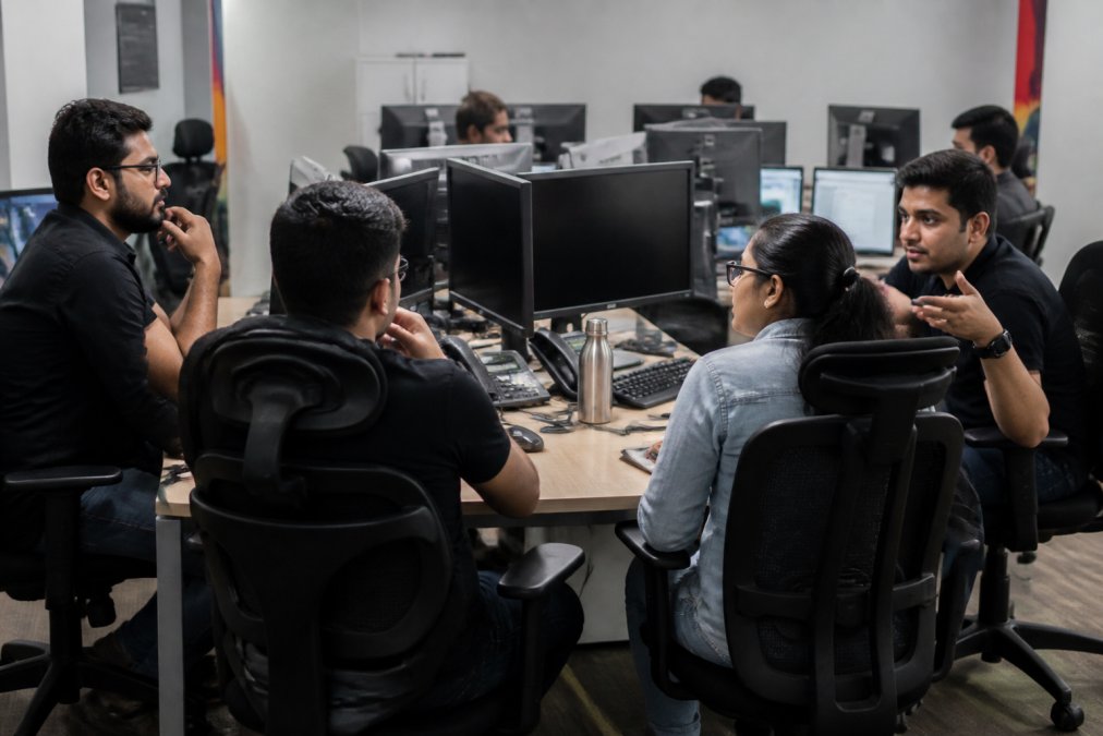 Security team in modern SOC environment collaborating at workstations, incident response playbooks visible on multiple monitors, analysts engaged in discussion around a central desk, professional lighting