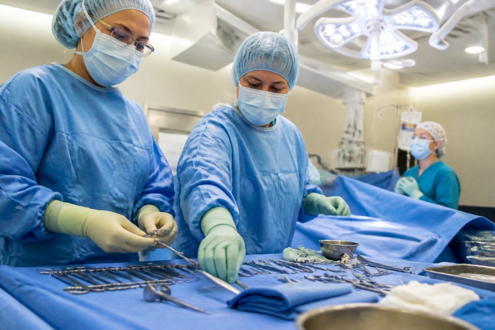 A scrubbed surgical team in full sterile gowns and gloves preparing instruments on a draped surgical table under bright overhead operating room lights, with a circulating nurse visible in the background