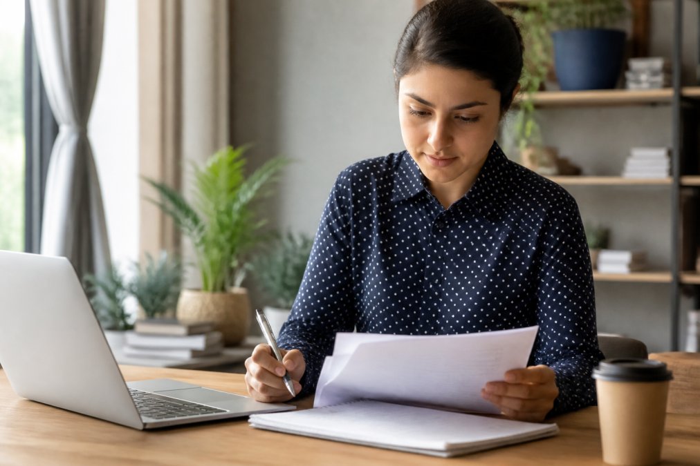 Entrepreneur working at a desk with a laptop, reviewing business formation documents and taking notes with a pen, natural office lighting from a window nearby
