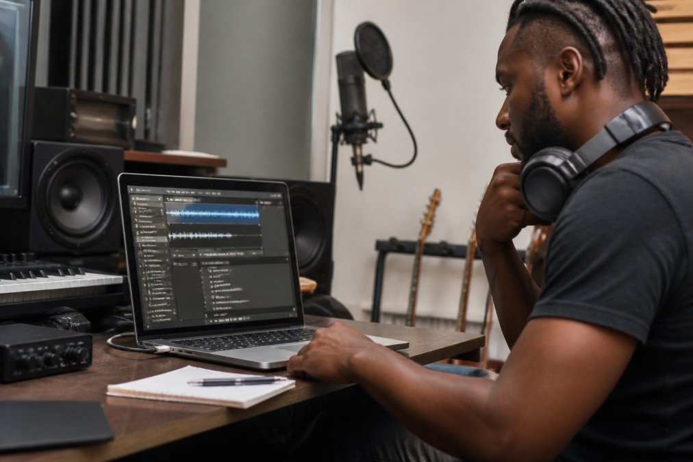 A musician at a desk in a home studio, looking at a laptop screen showing a digital audio workstation with metadata fields visible, headphones around the neck, and a microphone in the background