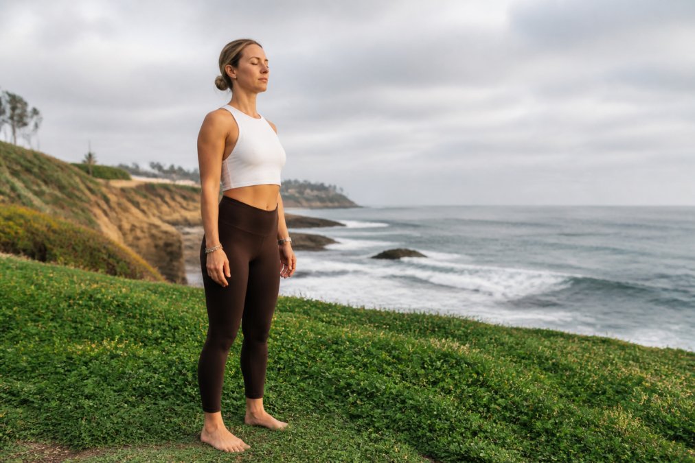 An active person doing a grounding exercise outdoors on grass or beach, barefoot, with relaxed posture and natural surroundings visible