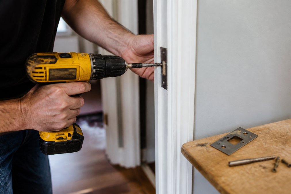 Hands using a drill to drive long screws into a metal strike plate on a wooden door jamb