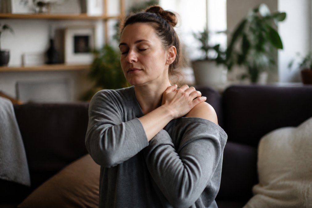 Person sitting comfortably indoors, gently placing hands on their shoulders and neck, demonstrating a somatic awareness and release technique with a calm, focused expression