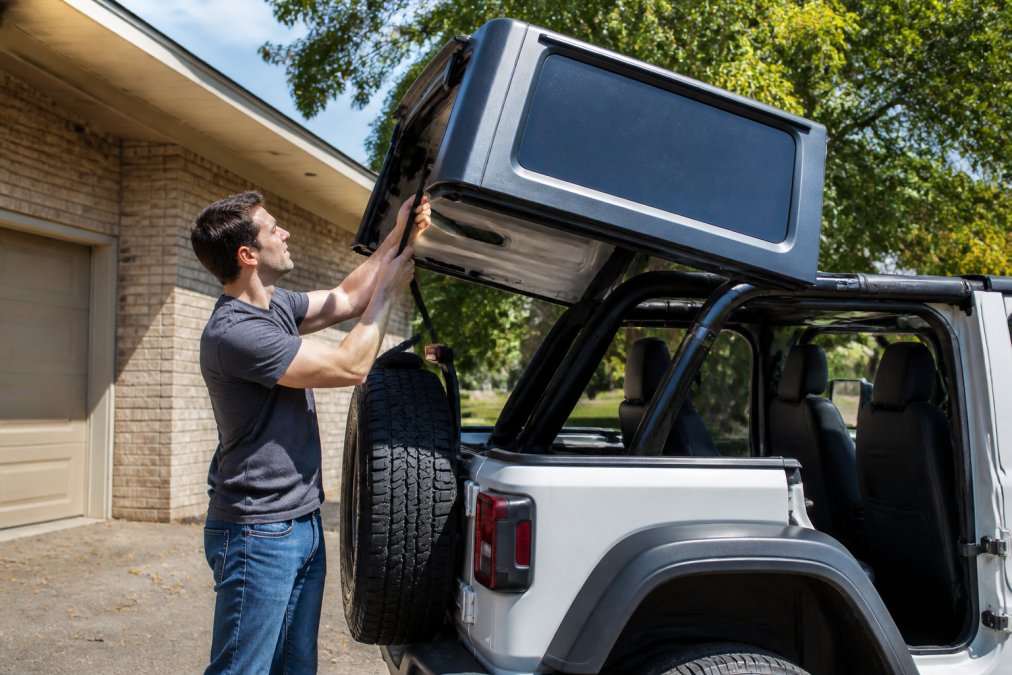 A single person using a padded lift assist strap to carefully lift the rear edge of a Jeep Wrangler hardtop, with the vehicle parked in a residential driveway on a sunny afternoon