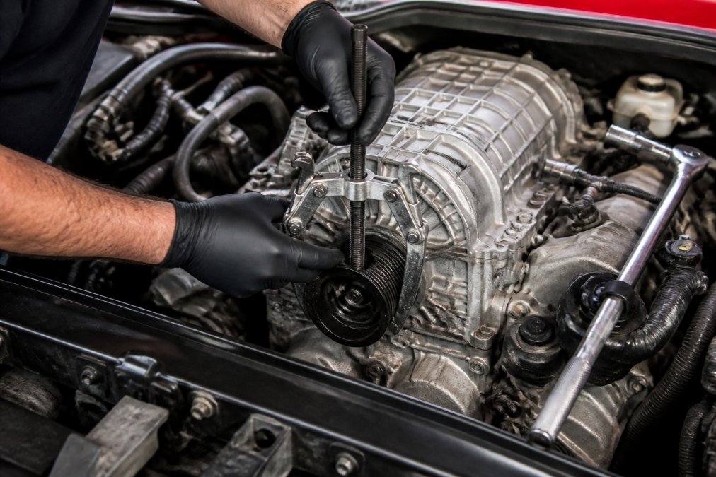 Close-up of a mechanic's gloved hands positioning a 3-jaw puller on a supercharger pulley in an open engine bay, a breaker bar resting on the intake manifold nearby, bright workshop lighting