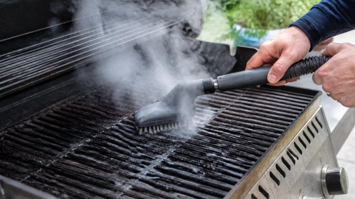 Close-up of hands scrubbing grill grates with a steam brush, steam rising visibly from the grates as baked-on grease lifts away