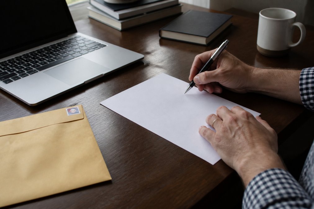 Close-up of hands writing a formal letter on white paper beside an open laptop and a stamped envelope ready to mail
