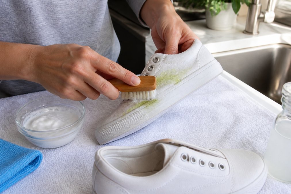 Close-up of hands scrubbing a white canvas sneaker with a soft-bristled brush and a white paste in a small bowl, set on a clean towel near a kitchen sink with bright natural lighting