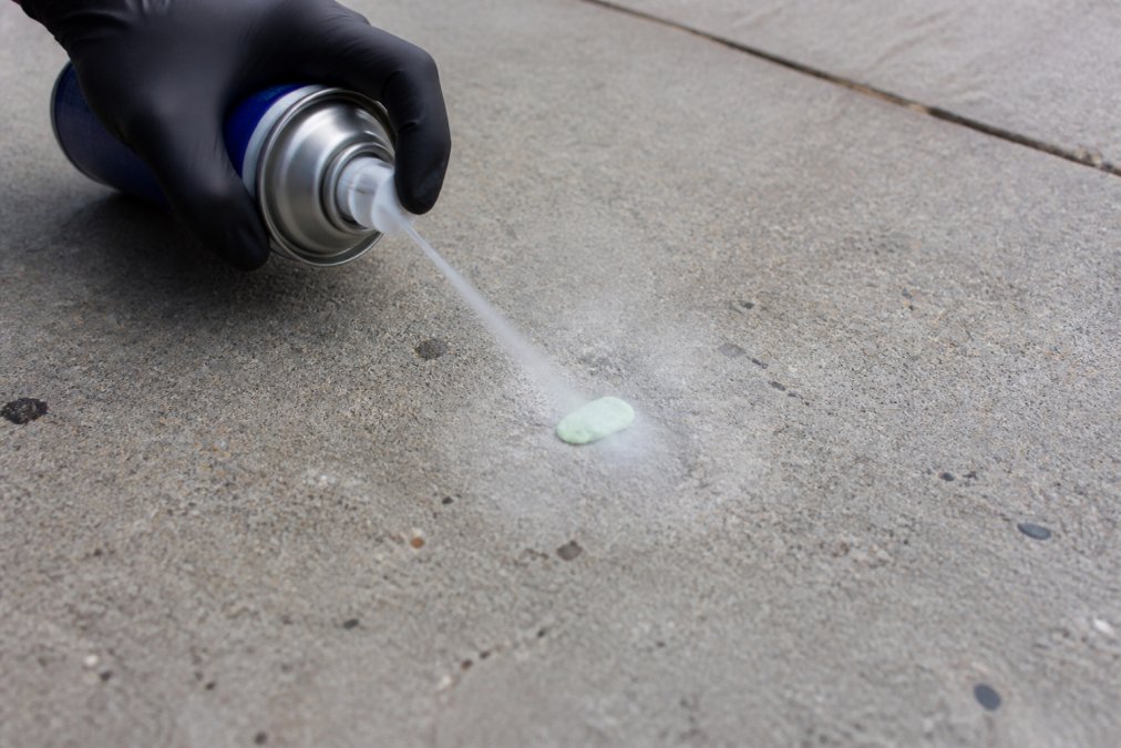 Close-up of a gloved hand aiming a can of compressed air at a piece of chewing gum stuck to a concrete sidewalk, with frosty vapor visible around the gum