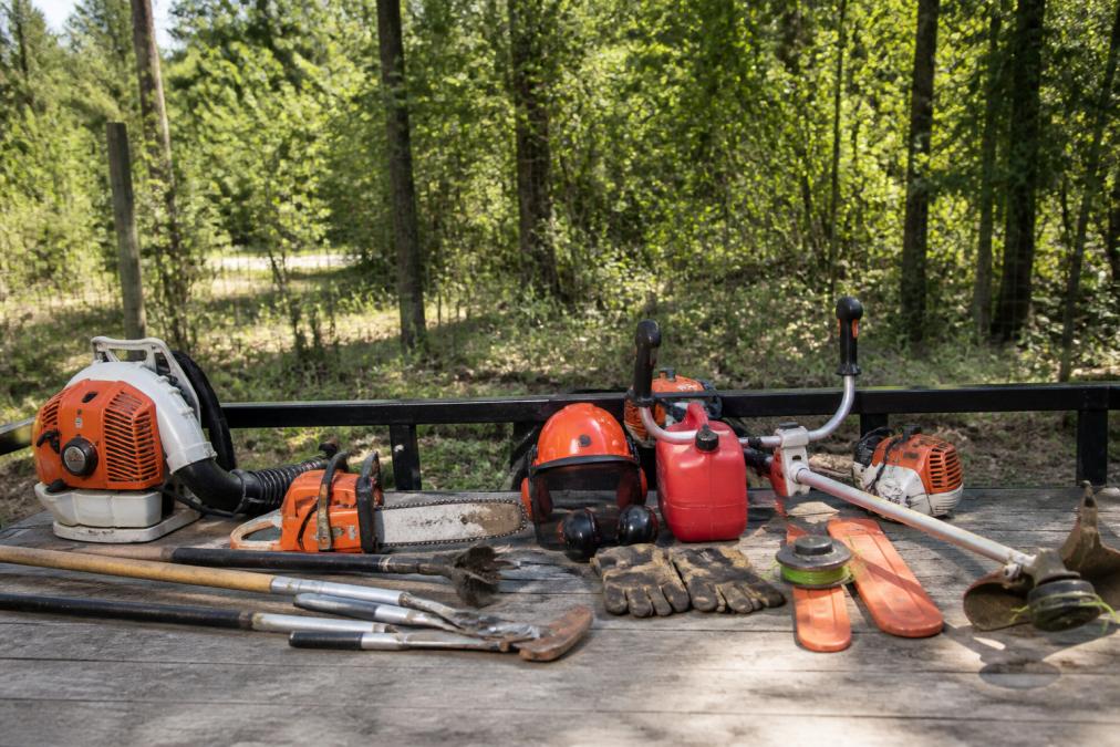 A worker in full protective gear including a cutting helmet and face shield operating a walk-behind brush mower on an overgrown wooded property, with thick saplings and tangled vines visible in the background, afternoon sunlight filtering through the tree canopy