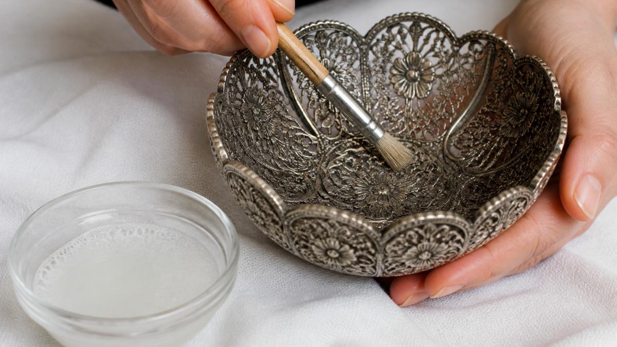 Close-up of hands using a soft-bristled brush to gently clean the recessed floral pattern of an ornate silver filigree bowl on a clean white cloth, with a small bowl of mild soapy water nearby