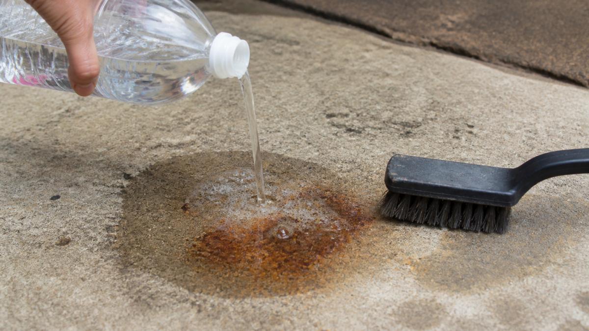 Close-up of white vinegar being poured onto a rust stain on concrete surface with a stiff-bristled brush nearby