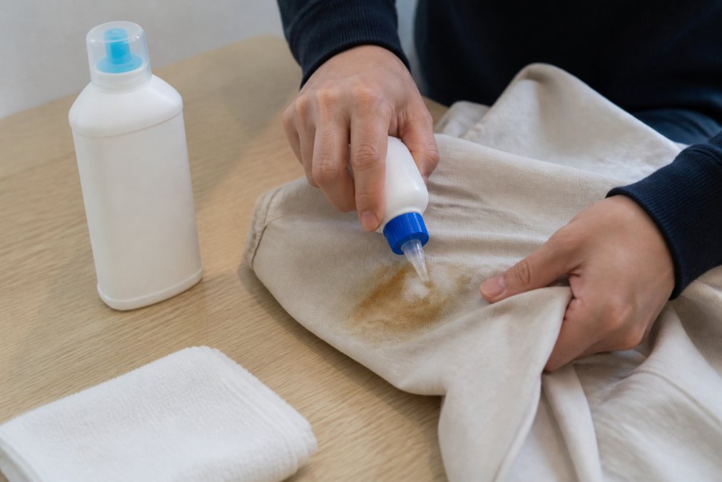 A person applying a surfactant-based stain remover to an oil stain on a garment before washing, with the product bottle visible