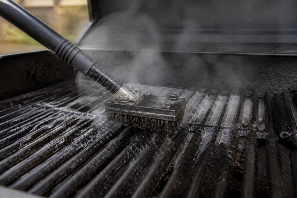 Close-up of steam brush in action on hot grill grates with visible steam rising and grease melting away, showing brush head making contact with carbonized buildup