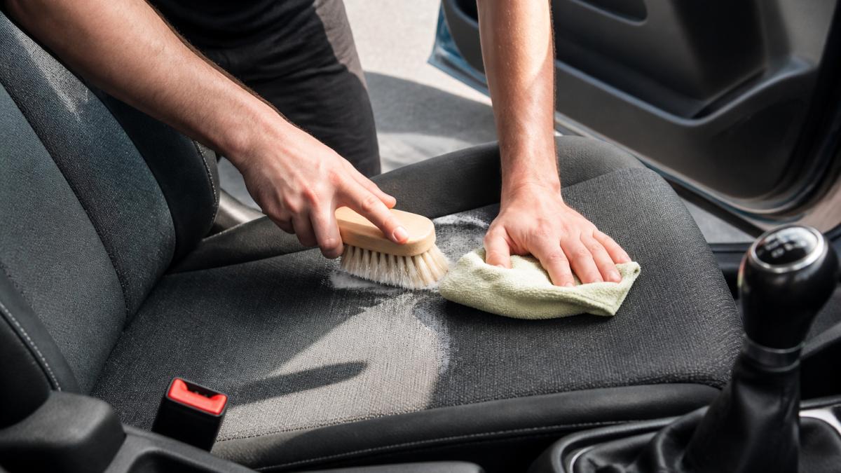 Close-up of hands using a soft brush and microfiber cloth to gently scrub a cloth car seat with cleaning solution, showing proper technique and lighting from the car window