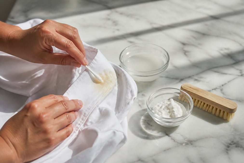 Close-up of hands applying hydrogen peroxide solution to yellow sweat stain on white dress shirt collar, with baking soda paste and soft brush visible on marble countertop, natural morning light