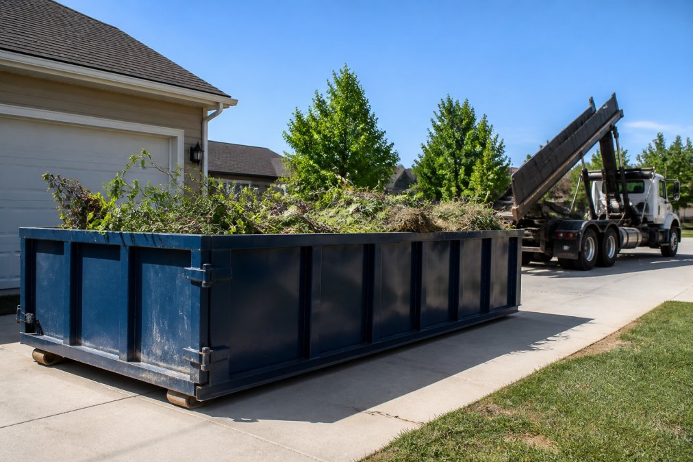 A roll off dumpster sitting on a residential driveway loaded with tree branches and grass clippings, with a truck in the background preparing for pickup on a clear day