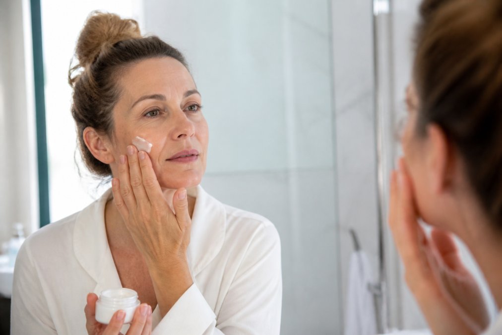A woman in her 40s with sensitive skin gently applying a small amount of healing balm to her cheek in a bright bathroom, soft natural light, calm expression