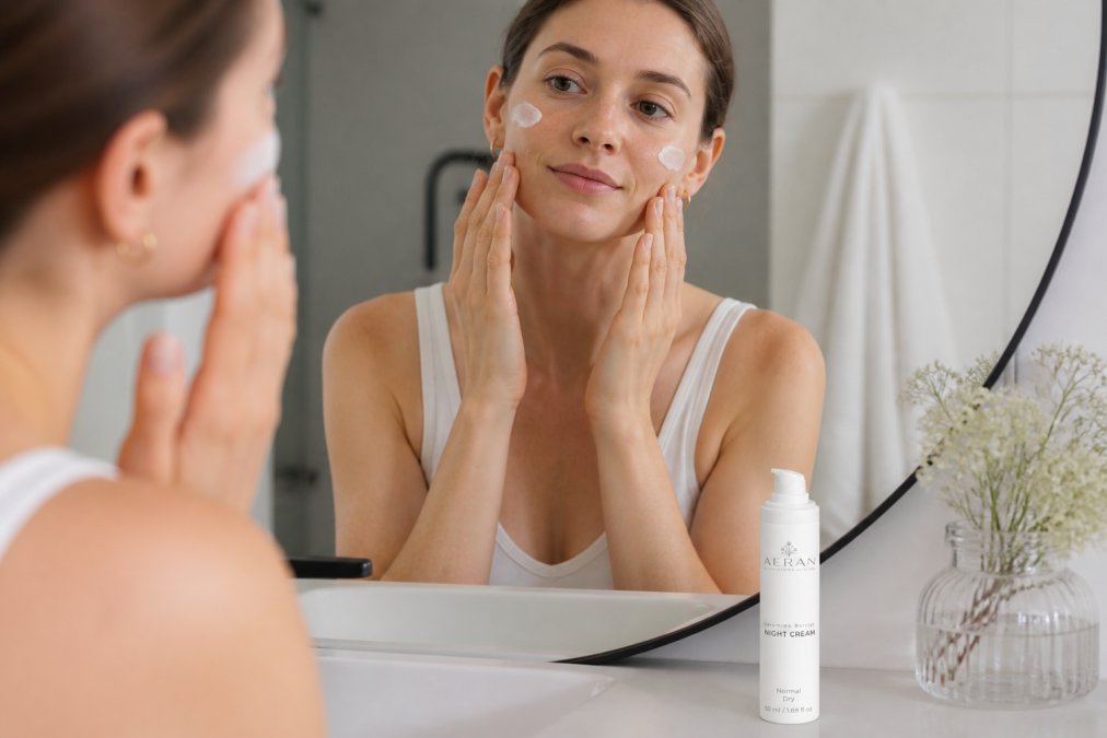 Person's hands applying barrier repair cream to face in front of bathroom mirror with soft warm lighting