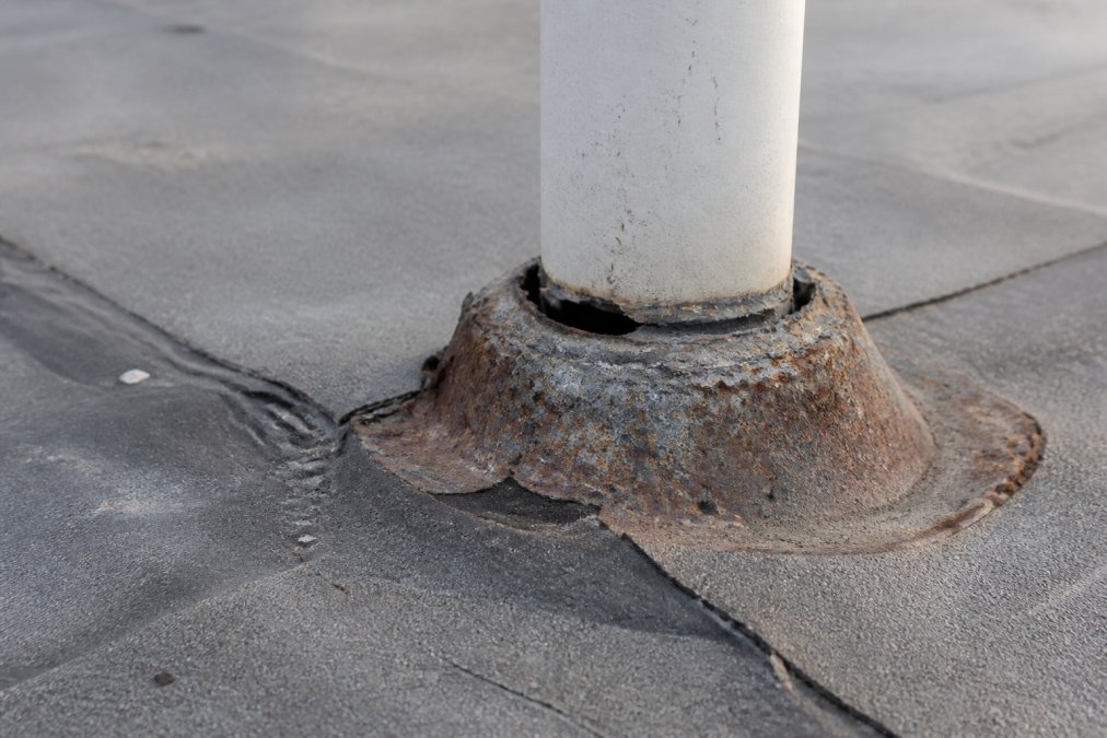 Close-up of deteriorated roof flashing showing rust, corrosion, and separation from the roof surface around a vent pipe penetration, with visible gaps and weathered sealant