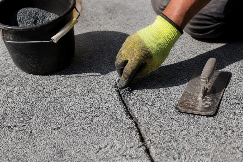 A close-up photo of a gloved hand pressing resin filler into a narrow crack on a grey resin-bound driveway, with a steel trowel resting nearby and soft daylight casting a shadow across the surface
