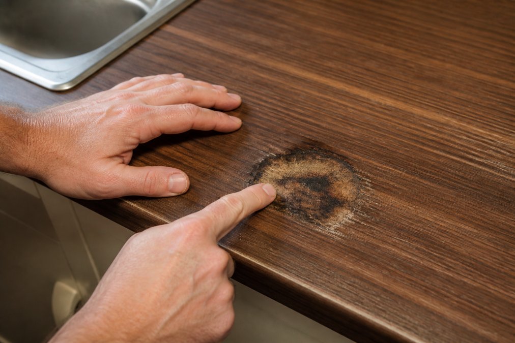 Close-up of hands carefully examining a deep burn mark on laminate worktop surface, showing the depth and extent of heat damage with natural kitchen lighting