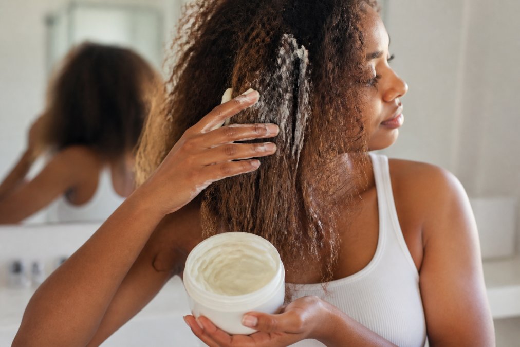 A person with visibly dry, frizzy hair applying a thick, creamy hair mask from a jar with their fingers in a bright bathroom setting