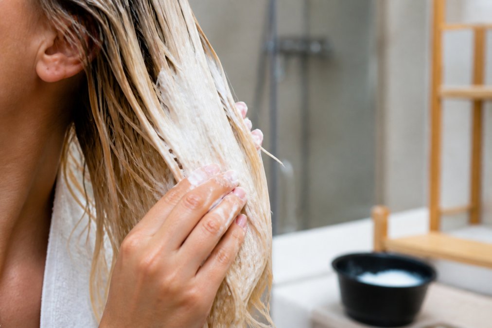 Close-up of a person's hand applying a thick, creamy hair mask to damp, bleached strands in a bright bathroom, product visibly coating each section of hair