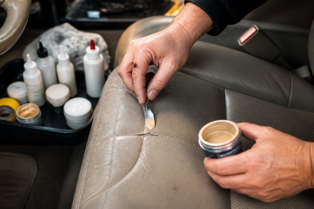 Close-up of a person's hands applying leather filler to a cracked car seat with a small spatula, in a well-lit garage setting