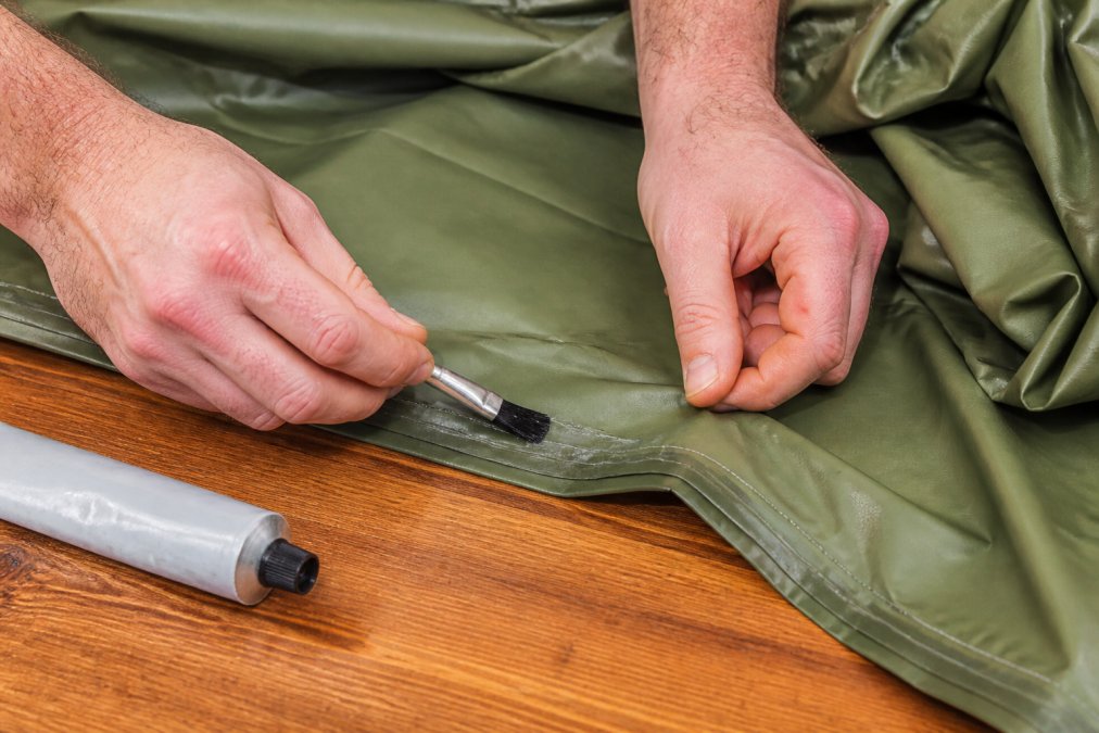 Close-up of hands applying seam sealer along the inside seam of a green waterproof poncho spread out on a wooden table, with a small brush and tube of sealant nearby