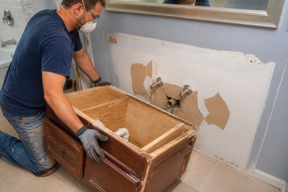 Contractor removing an old bathroom vanity to reveal wall plumbing and damaged drywall during the replacement process.