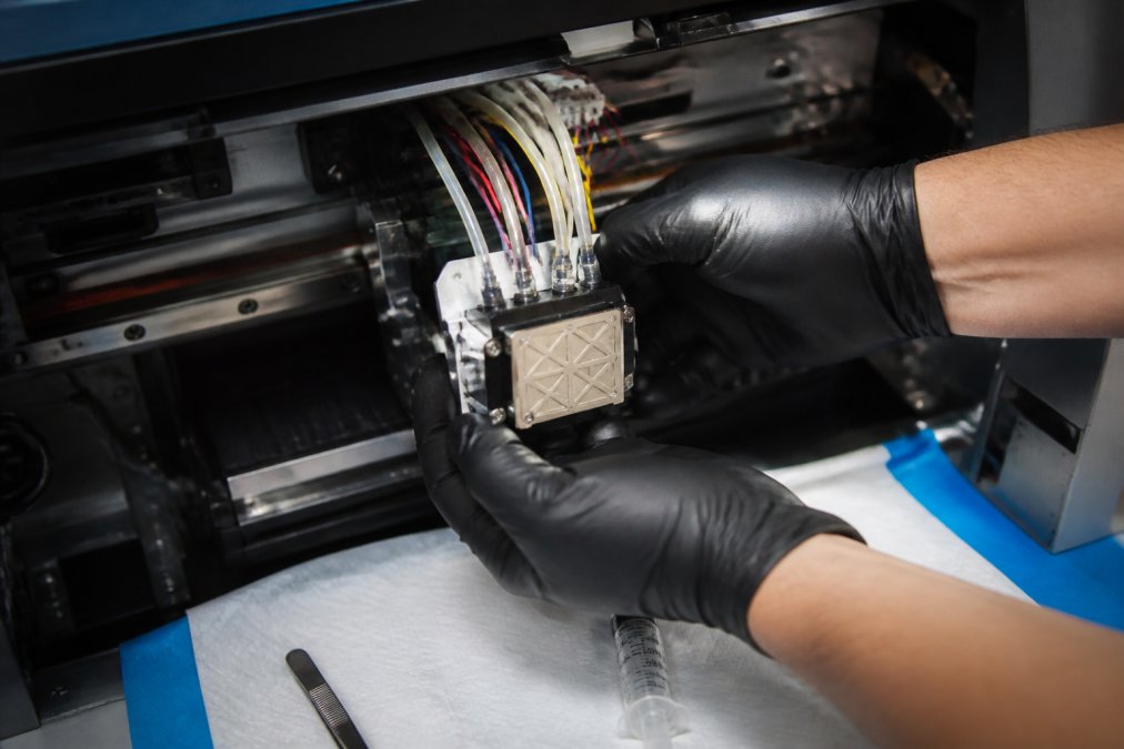 Close-up of a technician's gloved hands lifting a DTF printer printhead out of the carriage, with the printhead cable and ink lines visible and a clean work mat below