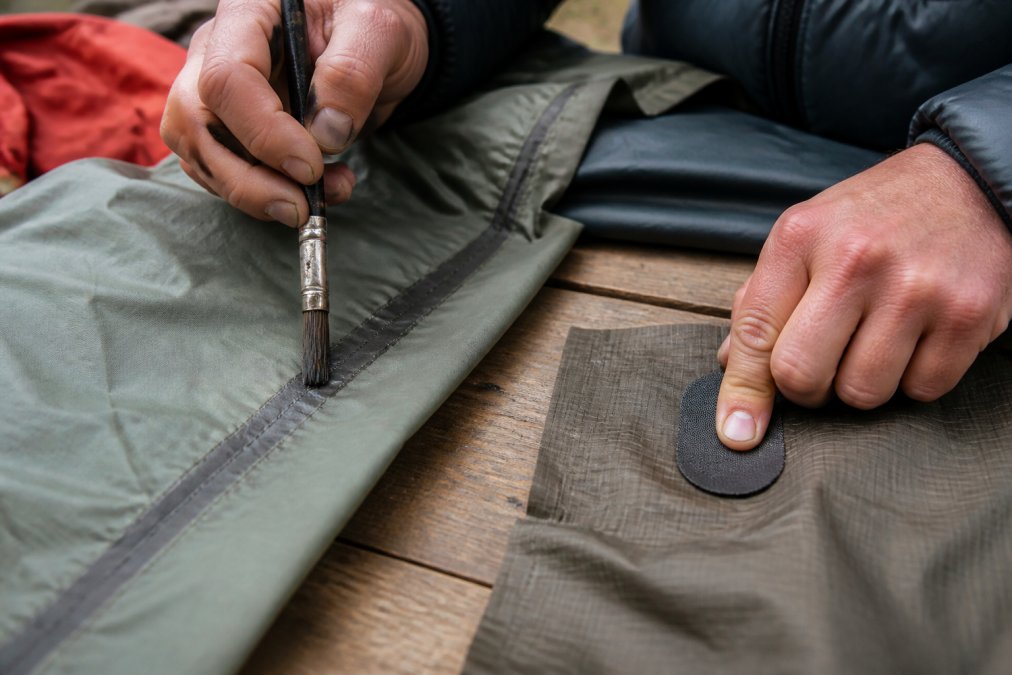 Hands applying seam sealer to a tent seam with a brush, and placing an adhesive fabric patch over a small tear in nylon material on a work surface